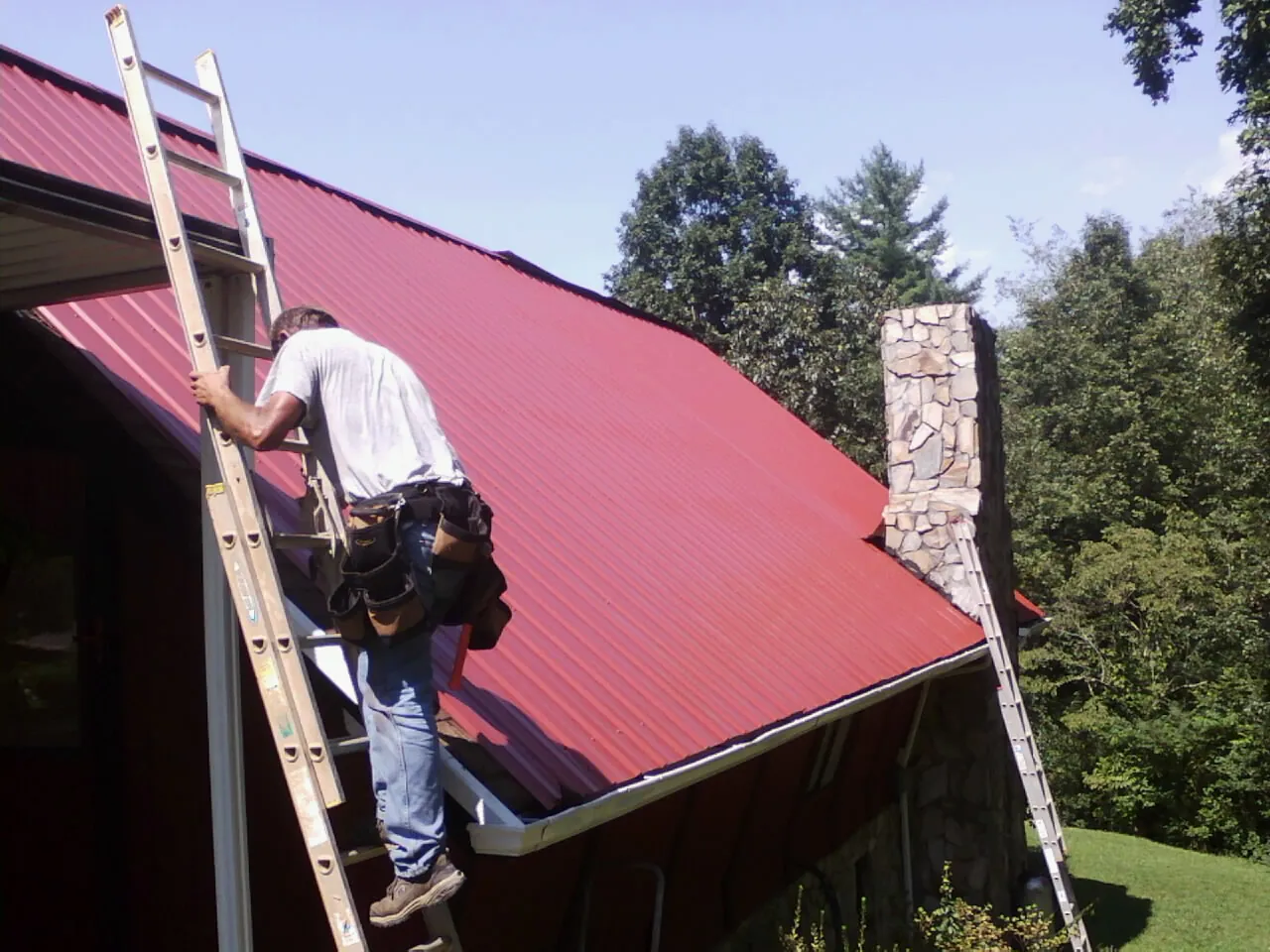 Roofer on a ladder beside a red metal roof and stone chimney — Soffit & Fascia Repair by Roofing Brothers in Cramerton